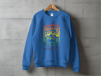 A blue sweatshirt by Garment Graphics hangs on a wooden hanger against a concrete wall, showcasing retro vibes with a colorful graphic that says Woodstock 1969, featuring floral elements and a guitar design celebrating the iconic festival.