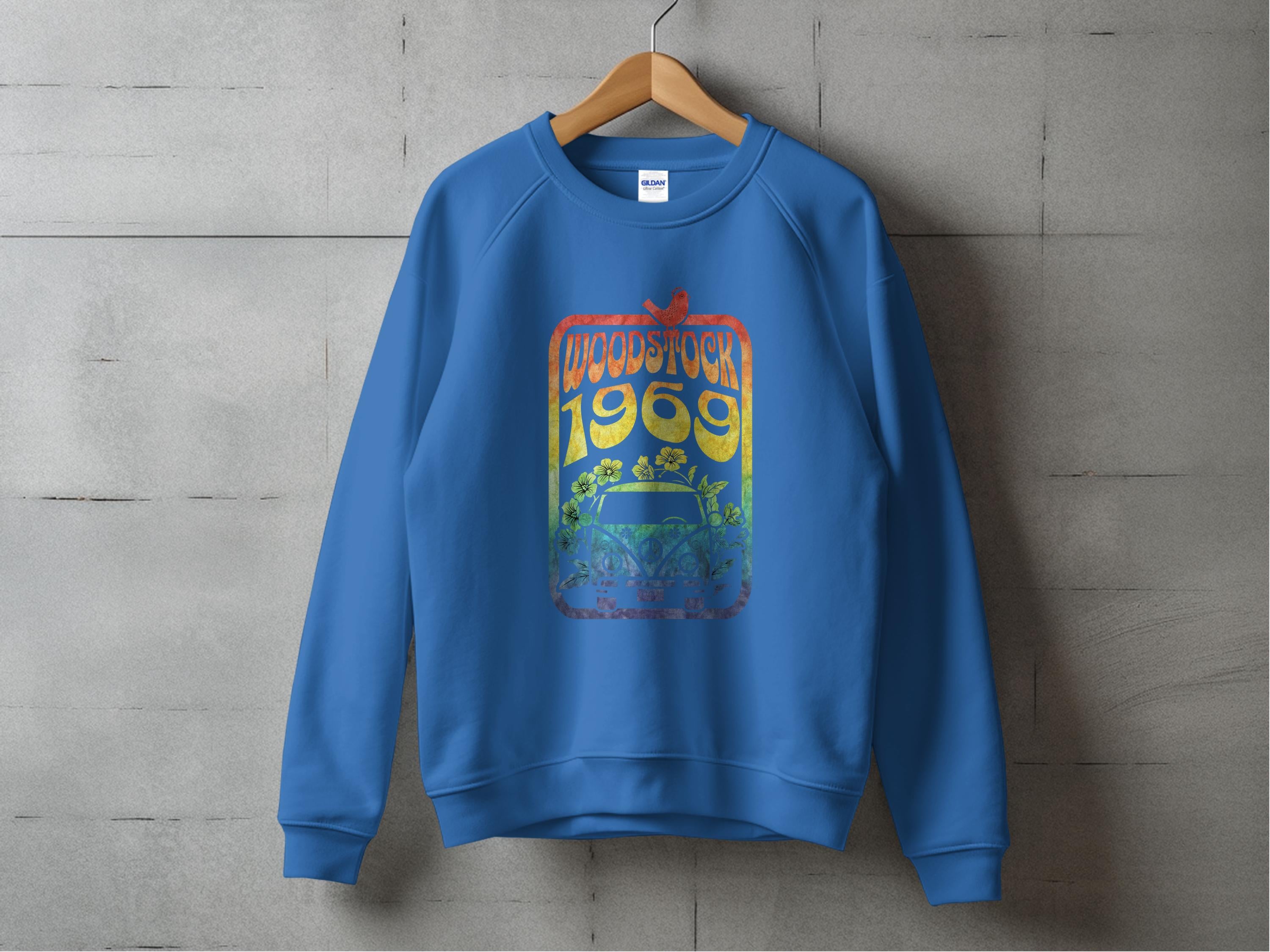 A blue sweatshirt by Garment Graphics hangs on a wooden hanger against a concrete wall, showcasing retro vibes with a colorful graphic that says Woodstock 1969, featuring floral elements and a guitar design celebrating the iconic festival.