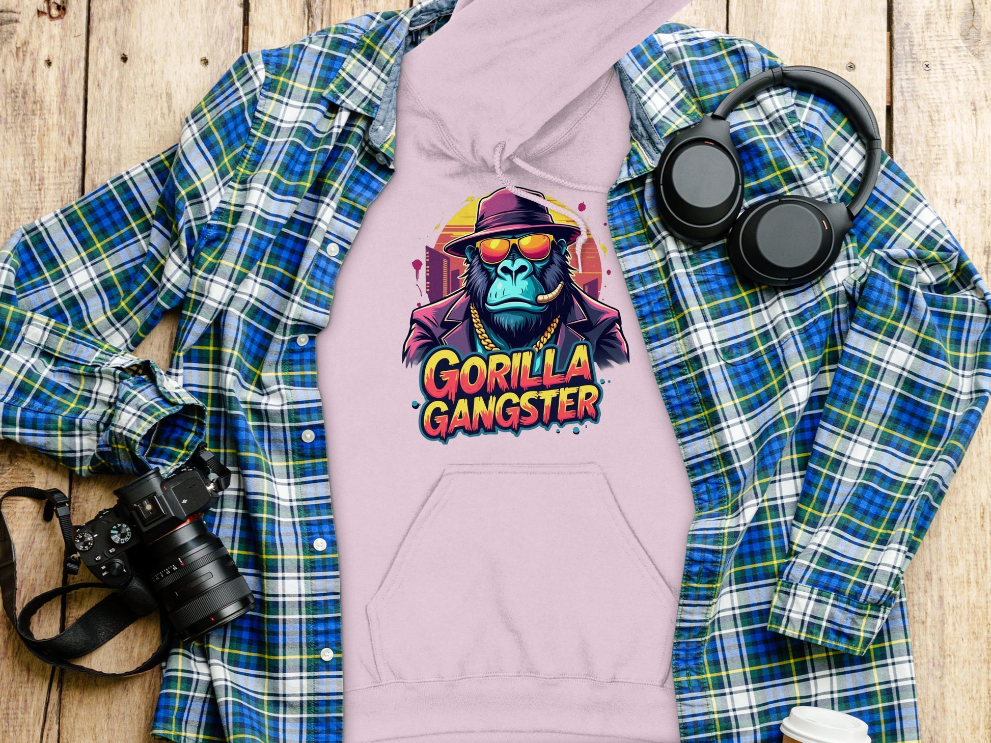 On a wooden surface lies a lavender hoodie from Garment Graphics featuring Gorilla Gangster artwork, paired with a blue and green plaid shirt. Completing the urban aesthetic are a camera, headphones, and a cup—ideal for streetwear enthusiasts.