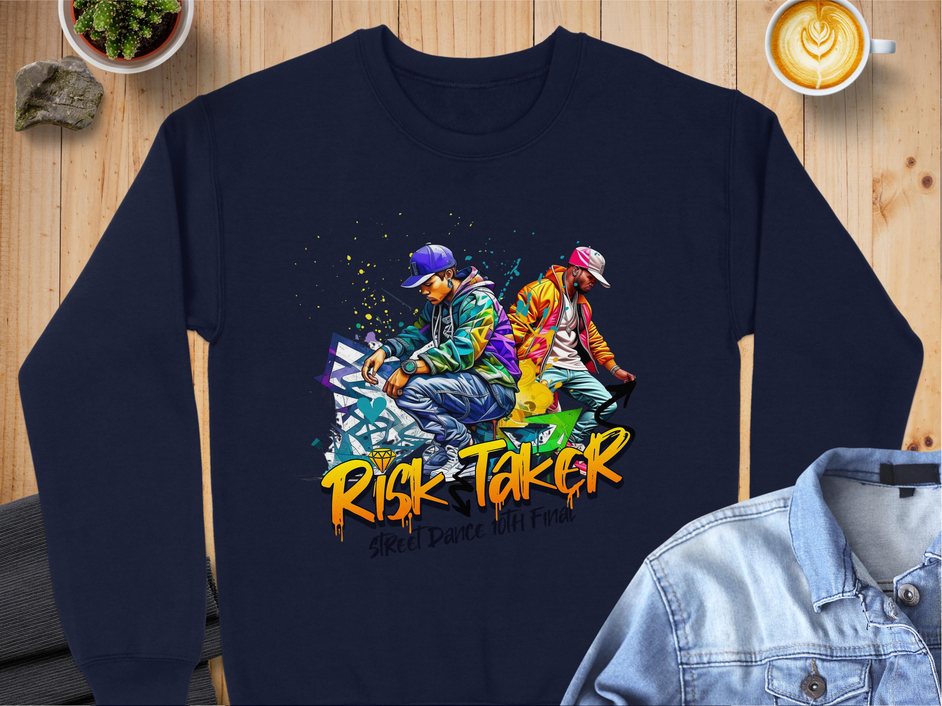 A navy blue sweatshirt from Garment Graphics features a vibrant graphic of two street dancers, capturing the essence of Risk Taker Street Dance with Street Dance Unit Fire below. Its styled with a denim jacket, coffee cup, and small plants on a wooden surface.