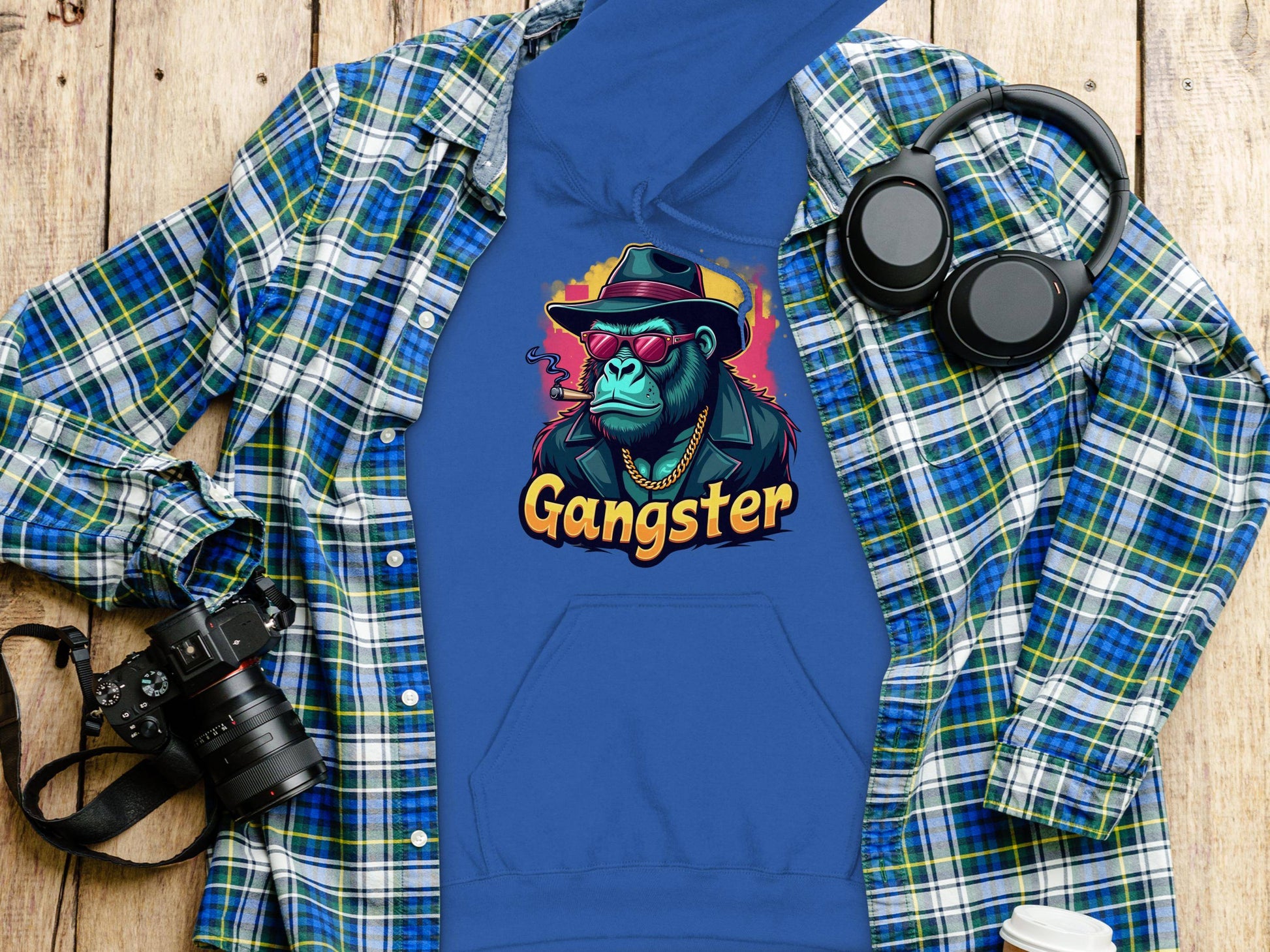 A blue hoodie by Garment Graphics in medium-heavy fabric features a Gangster gorilla graphic with sunglasses, a hat, and gold chain. Styled with a green-blue plaid shirt, camera, headphones, and coffee cup on wooden surface for a smoky, cool look.