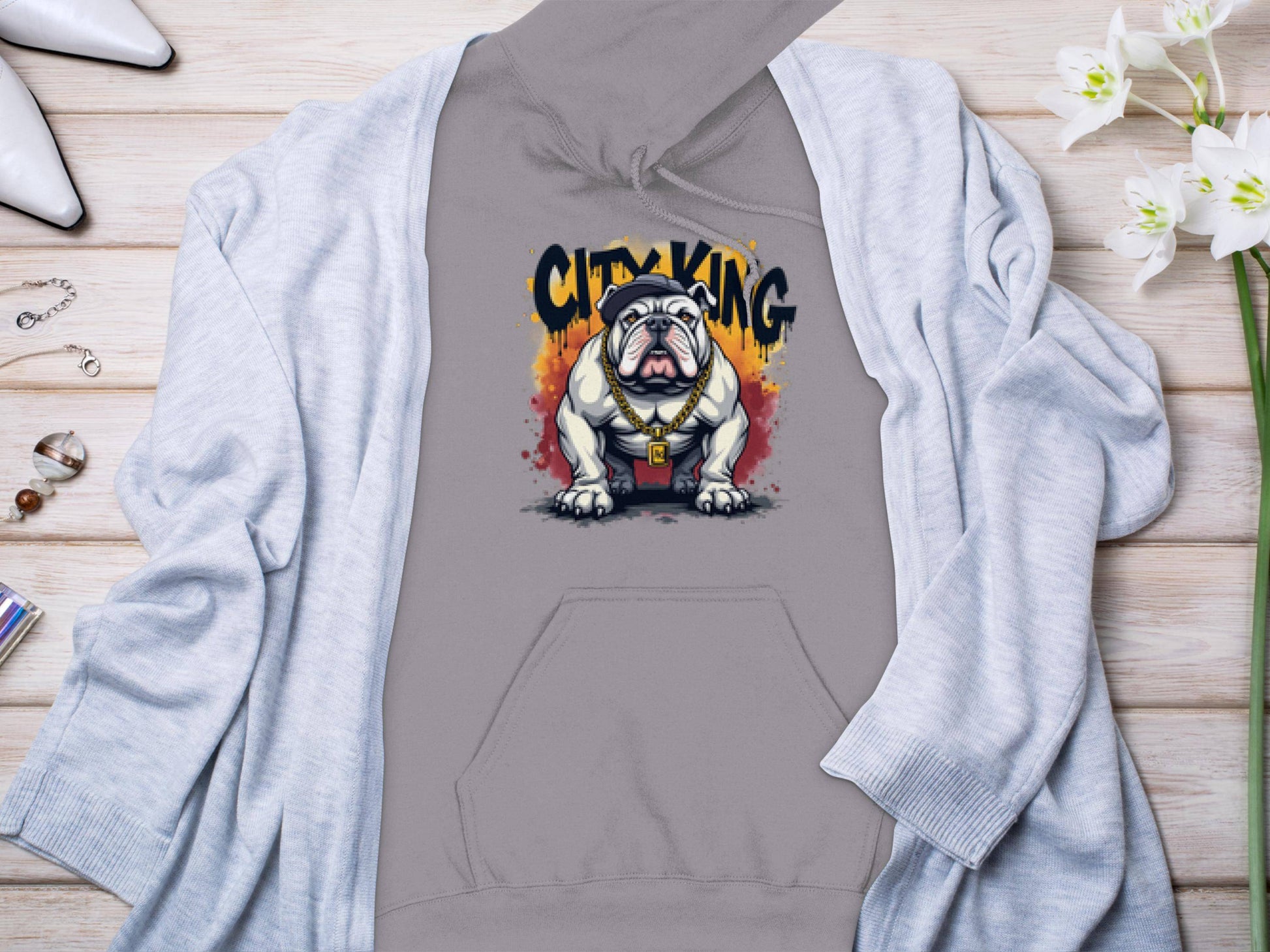 The Garment Graphics City King gray hoodie, featuring a graphic of a bulldog with a gold collar, is laid on a light wooden surface. Its complemented by a gray cardigan, white high heels, watch, ring, and white flowers—ideal for casual wear.