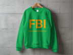 A green sweatshirt by Garment Graphics, with unisex sizing, hangs on a wooden hanger against a gray concrete wall. It features bold yellow text reading FBI Female Boob Inspector, adding a humorous twist to the classic design.