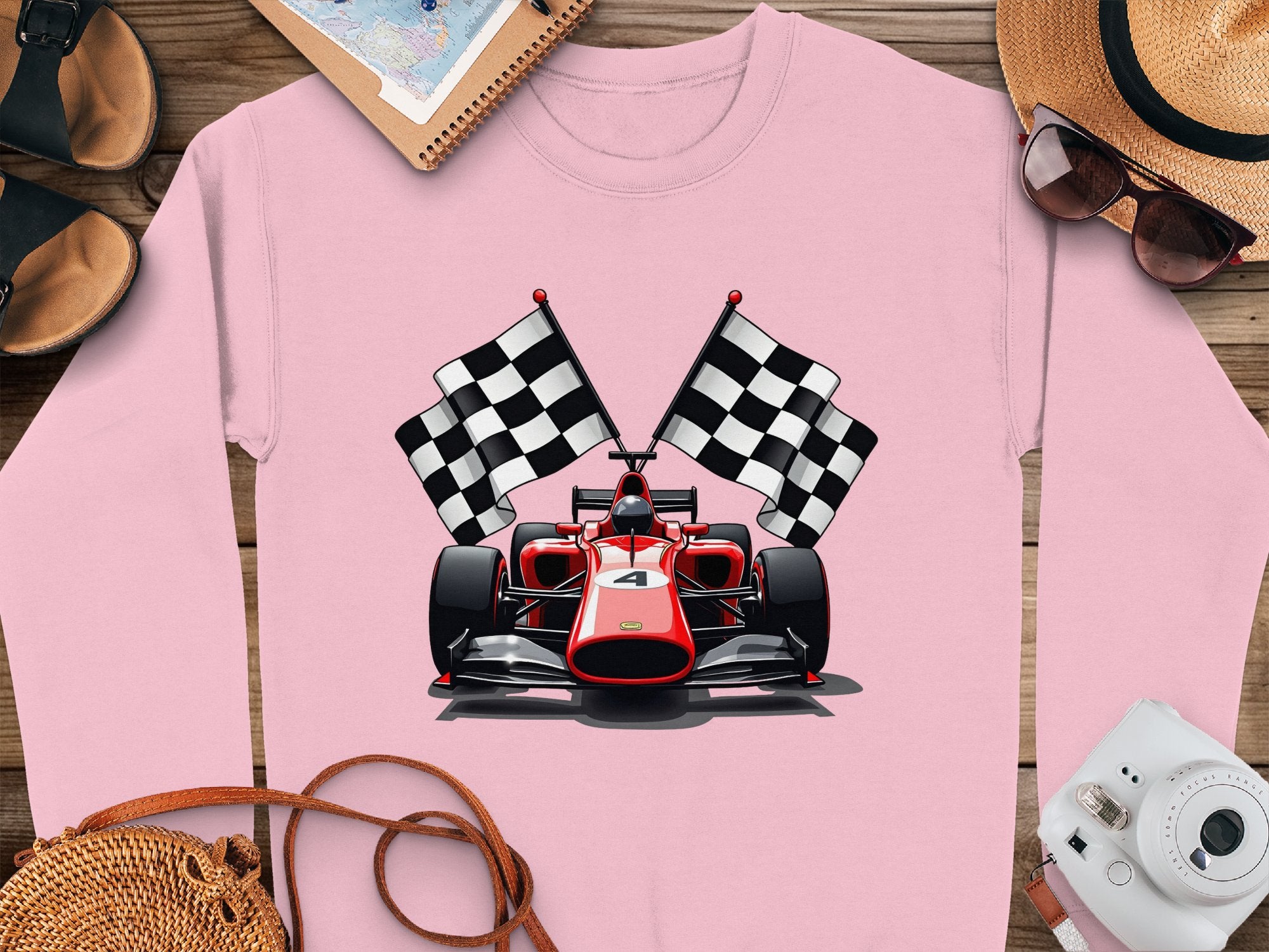 A vibrant pink Garment Graphics sweatshirt features a red racing car with checkered flags design, ideal for motorsport fans. Nearby on the wooden surface are a camera, sunhat, sunglasses, sandals, and a notebook.