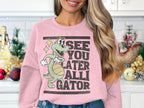 In a festive kitchen, a woman wears a pink Garment Graphics sweatshirt with a fun alligator graphic. The green cartoon alligator holds a basketball among stars, accompanied by the playful text SEE YOU LATER ALLIGATOR.