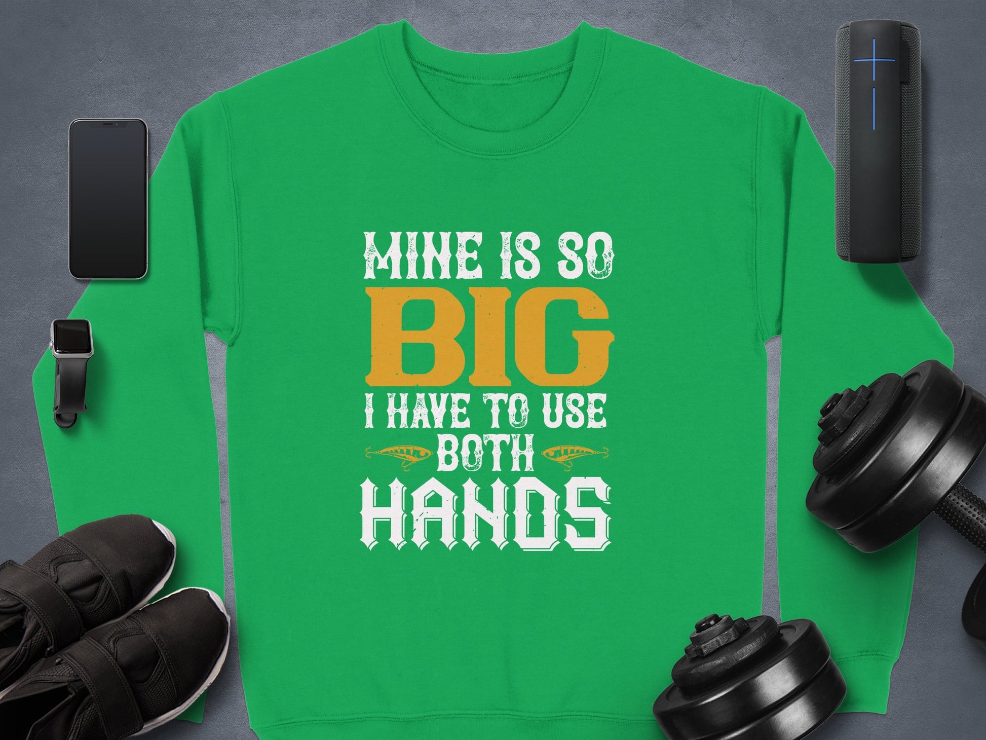 Garment Graphics bold statement green sweatshirt features white and orange text: Mine is so big I have to use both hands. Displayed with a smartphone, smartwatch, speaker, black shoes, dumbbells, and a cylindrical object on a gray backdrop. Ideal for unique sweatshirt enthusiasts.