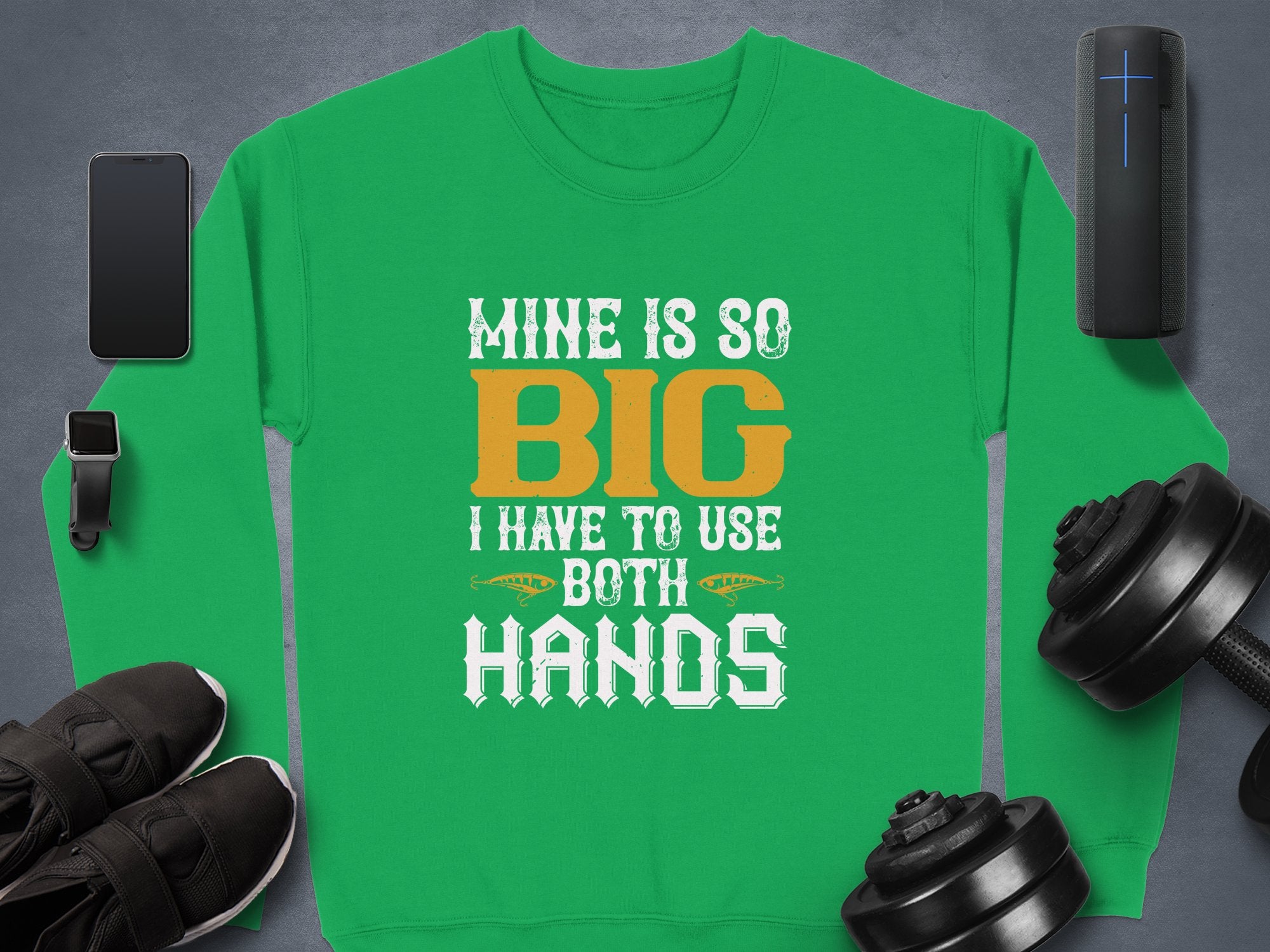 Garment Graphics bold statement green sweatshirt features white and orange text: Mine is so big I have to use both hands. Displayed with a smartphone, smartwatch, speaker, black shoes, dumbbells, and a cylindrical object on a gray backdrop. Ideal for unique sweatshirt enthusiasts.