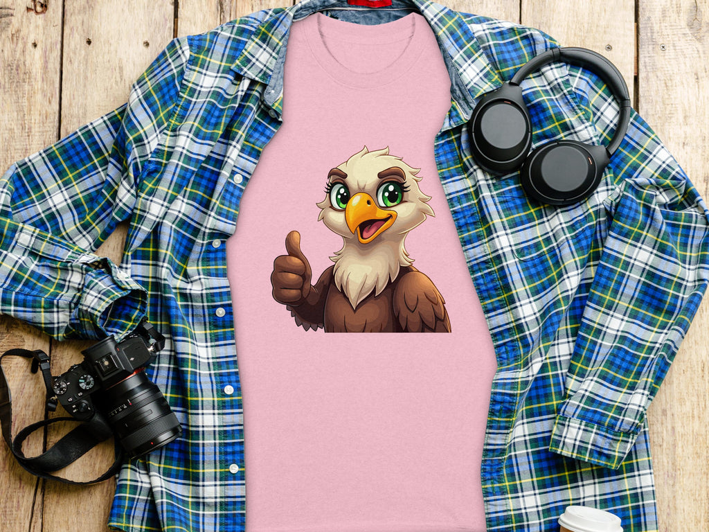 The Garment Graphics Eagle T-shirt, ideal for a casual look, has a cartoon eagle giving a thumbs up on pink fabric. Its styled amidst essentials like a blue and green plaid shirt, headphones, a camera, and a takeaway coffee cup for that classic fit vibe on wood.
