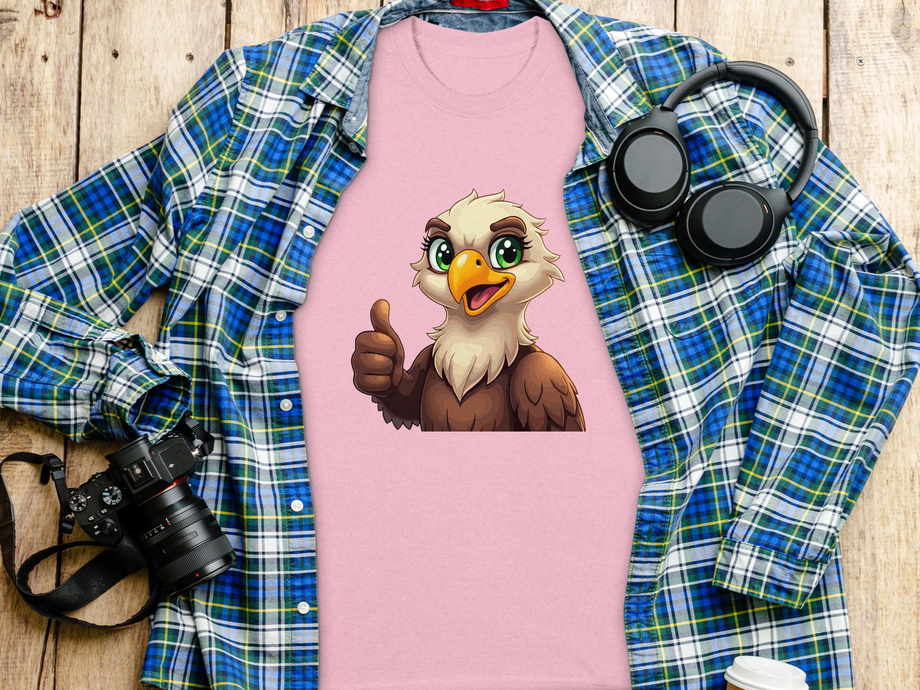 The Garment Graphics Eagle T-shirt, ideal for a casual look, has a cartoon eagle giving a thumbs up on pink fabric. Its styled amidst essentials like a blue and green plaid shirt, headphones, a camera, and a takeaway coffee cup for that classic fit vibe on wood.