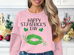 In a kitchen adorned with festive decor, a woman wears Garment Graphics pink sweatshirt featuring shamrocks, Happy St. Patricks Day greeting, and a green lip print capturing the clover kiss vibe.