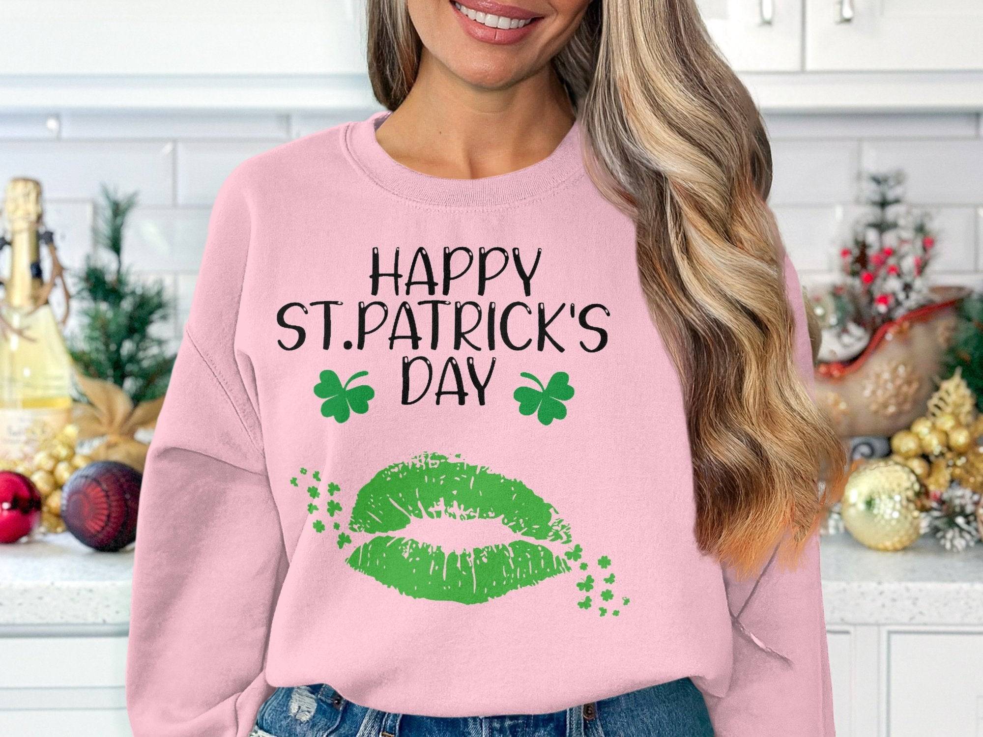 In a kitchen adorned with festive decor, a woman wears Garment Graphics pink sweatshirt featuring shamrocks, Happy St. Patricks Day greeting, and a green lip print capturing the clover kiss vibe.