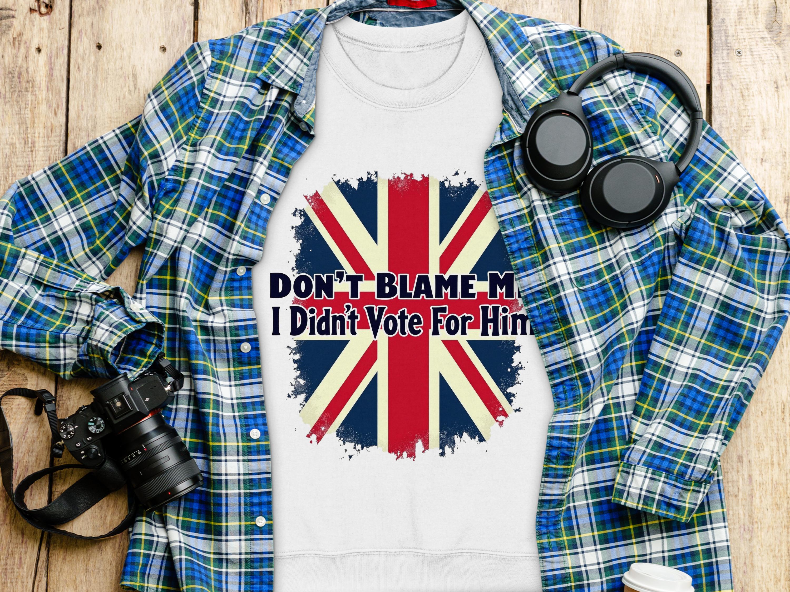 A Garment Graphics sweatshirt featuring a bold Union Jack and the text Dont Blame Me, I Didnt Vote For Him is displayed on wood, accompanied by a blue plaid shirt, headphones, a camera, and a water bottle.