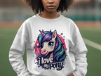 A person is wearing a Garment Graphics white sweatshirt featuring a colorful cartoon pony-unicorn with a flowing mane and the text I love my pony. The background shows blurred outdoor scenery.