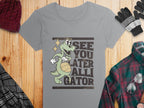 Garment Graphics Gray Alligator T-Shirt for women features a cartoon alligator holding a basketball with the phrase See you later alligator in bold. Styled with a red knit hat, gray gloves, plaid jacket, and glasses on a wooden surface.