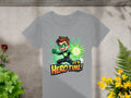 Garment Graphics presents a gray t-shirt featuring a young hero with brown hair and a green superhero design. The character poses dynamically with a glowing green orb, encircled by vibrant green and yellow plants, declaring, Its Hero Time! Ideal for any colorful wardrobe.