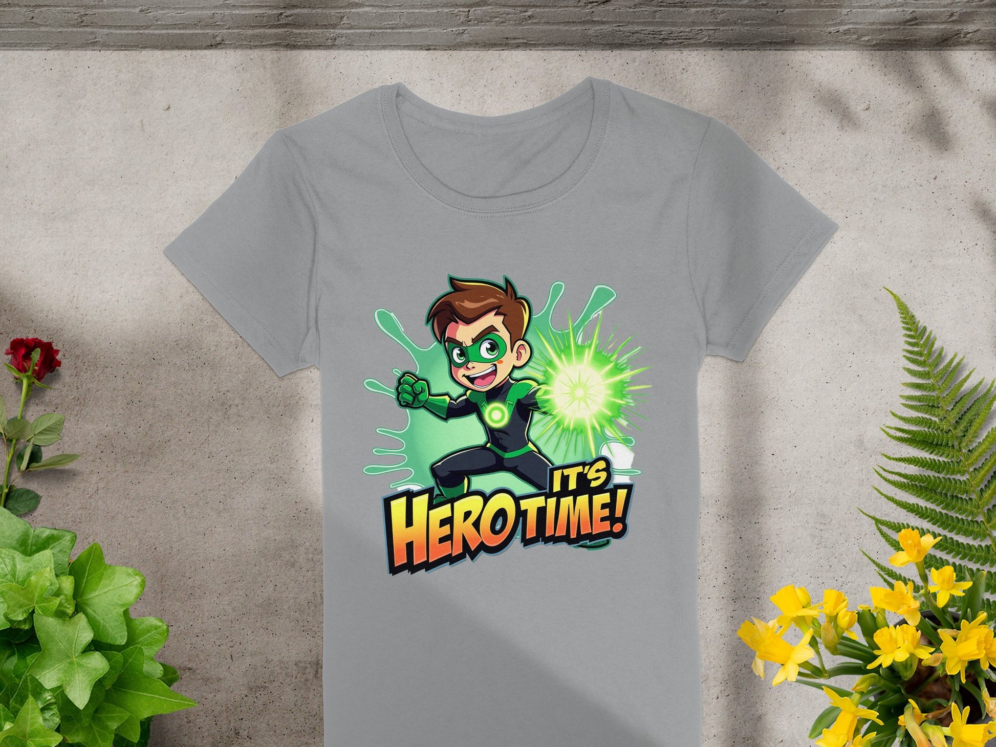 Garment Graphics presents a gray t-shirt featuring a young hero with brown hair and a green superhero design. The character poses dynamically with a glowing green orb, encircled by vibrant green and yellow plants, declaring, Its Hero Time! Ideal for any colorful wardrobe.