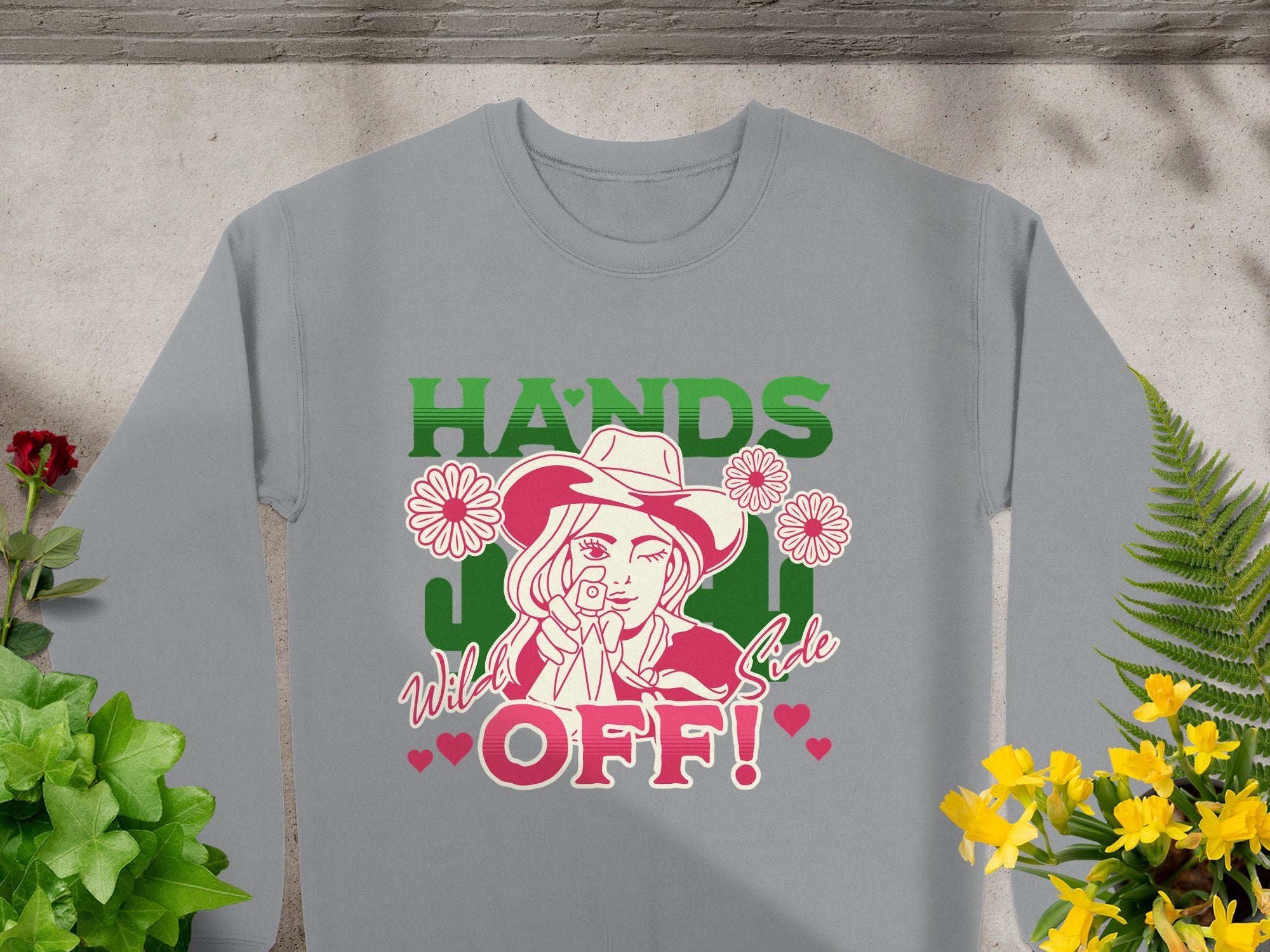 The Garment Graphics Hands Off Wild Side Sweatshirt features a line drawing of a person in a cowboy hat, with bold HANDS OFF! in green and pink. It is adorned with pink daisies, red hearts, greenery, and yellow flowers for added flair in a loose-fit style.