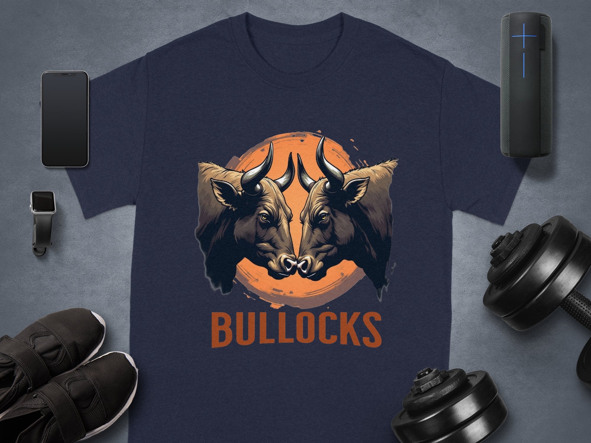 A navy blue Garment Graphics Pintable T-Shirt shows two bulls facing an orange circle with BULLOCKS below. Ideal for tech fans, this classic fit supports family sizing and is bordered by a smartphone, smartwatch, earphones, speaker, shoes, and dumbbells.