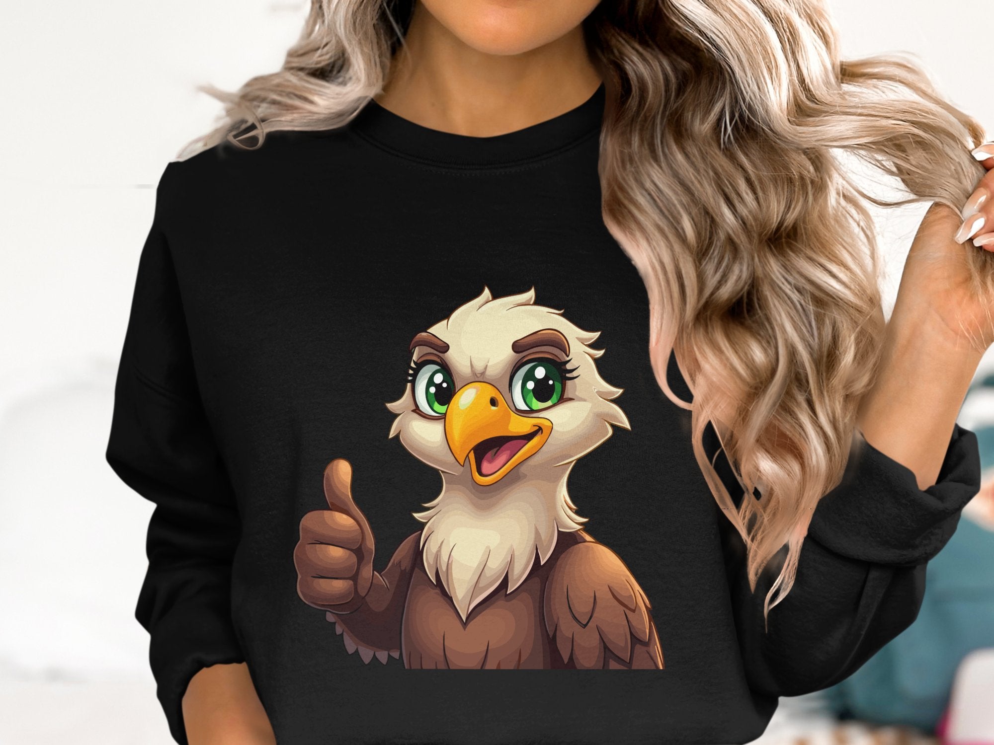 A person with long, wavy blonde hair sports a black Garment Graphics sweatshirt featuring a cartoon eagle giving a thumbs-up, adding flair to the simple light-colored interior backdrop.
