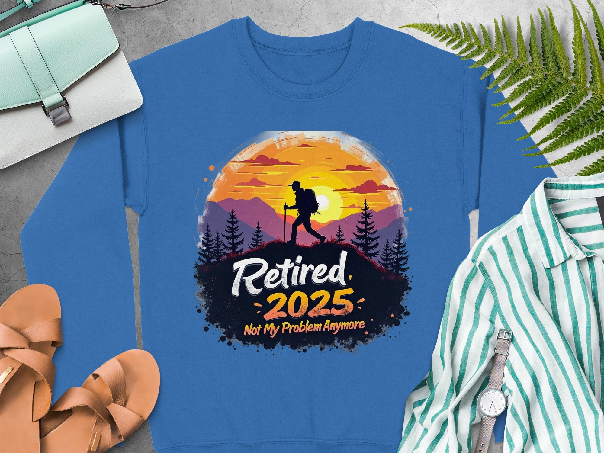 Garment Graphics presents a blue retirement sweatshirt featuring a hiker silhouette at sunset with trees. The Retired 2025, Not My Problem Anymore phrase adds charm, ideal for unisex sizing. Pair it with a purse, fern, striped shirt, shoes, and watch for a complete look.