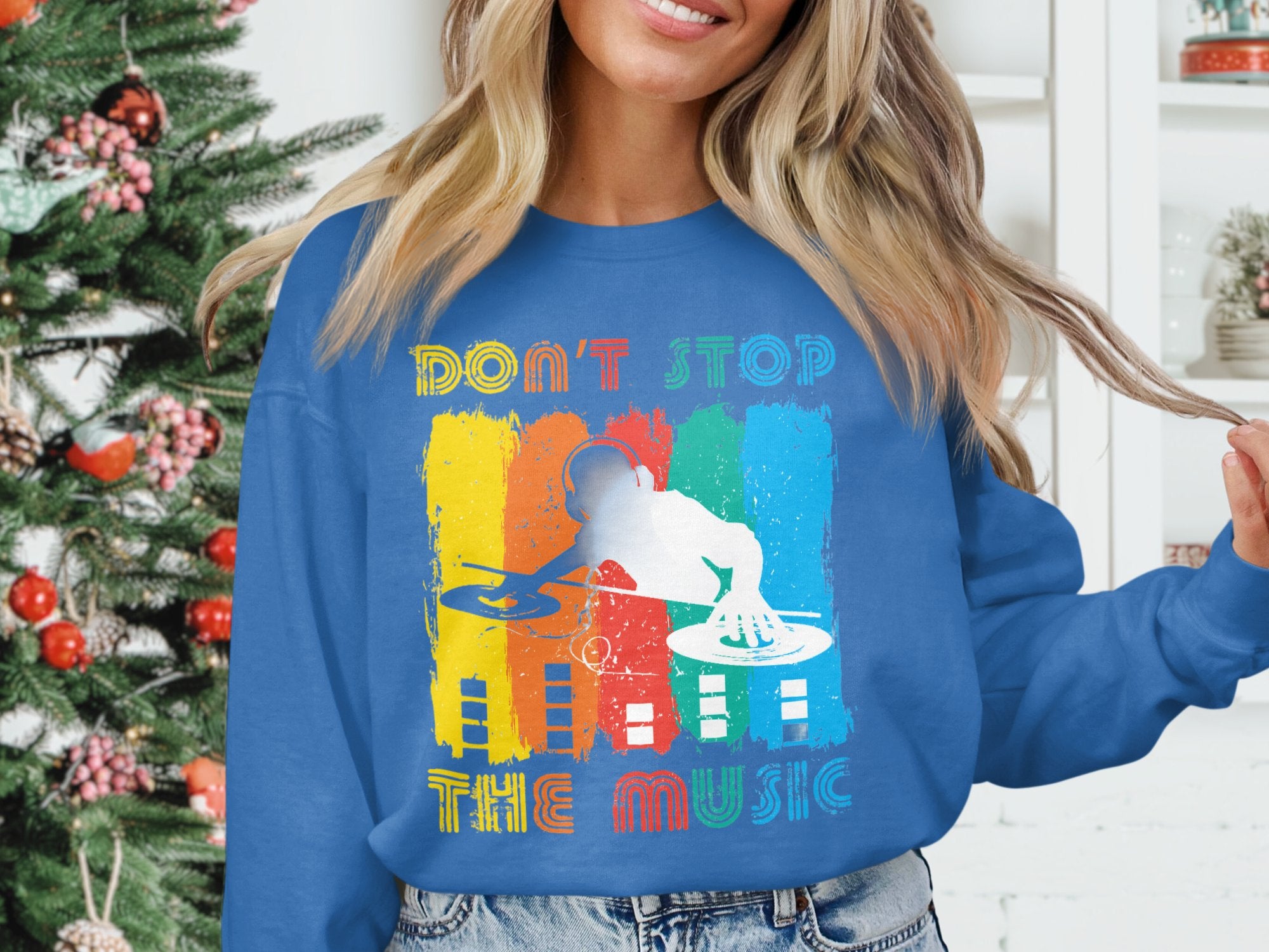 Indoors, a person stands near a decorated Christmas tree wearing Garment Graphics blue sweatshirt featuring a vibrant DJ design and the text Dont Stop the Music. Ideal for music lovers, shelves with small decorations appear in the background.