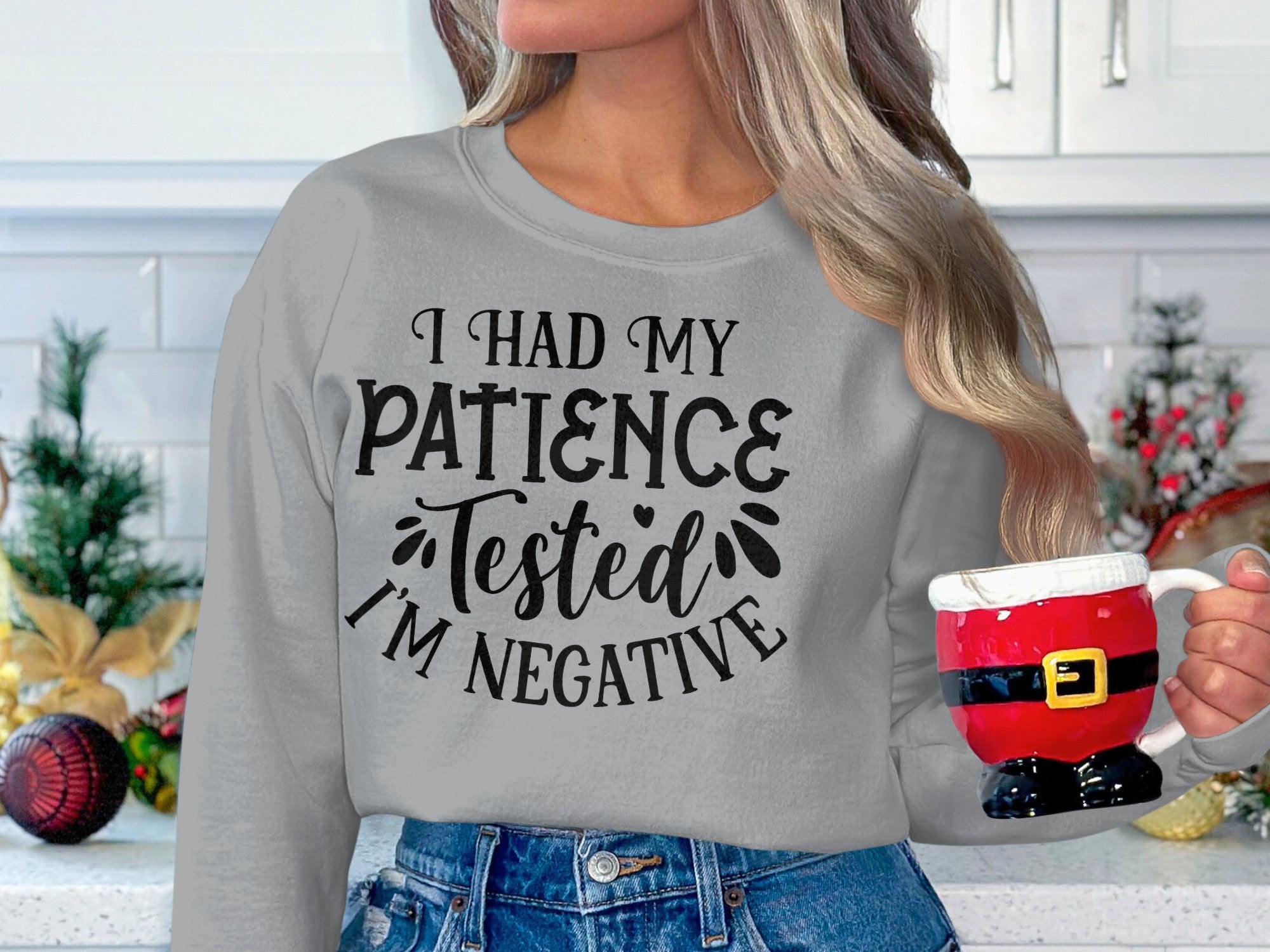 A person with long hair holds a Santa Claus-themed mug, wearing a Garment Graphics sweatshirt with the text, I had my patience tested Im negative. The cozy scene in a white kitchen filled with holiday decor is ideal for casual festive outings.