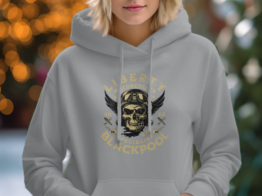 A white hoodie featuring a winged skull and Liberty Motorcycles Specialist Blackpool 2023 by Garment Graphics sits on wood, surrounded by a hat, sunglasses, sandals, camera, notebook, and woven bag—ideal companions for the iconic Blackpool hoodies style.