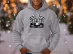 A person in a stylish Garment Graphics gray hoodie adorned with a racing car and checkered flags stands outside with hands in pockets. The backdrop includes blurred trees, lights, and falling snowflakes, capturing the essence of motorsport enthusiasts on a wintery day.