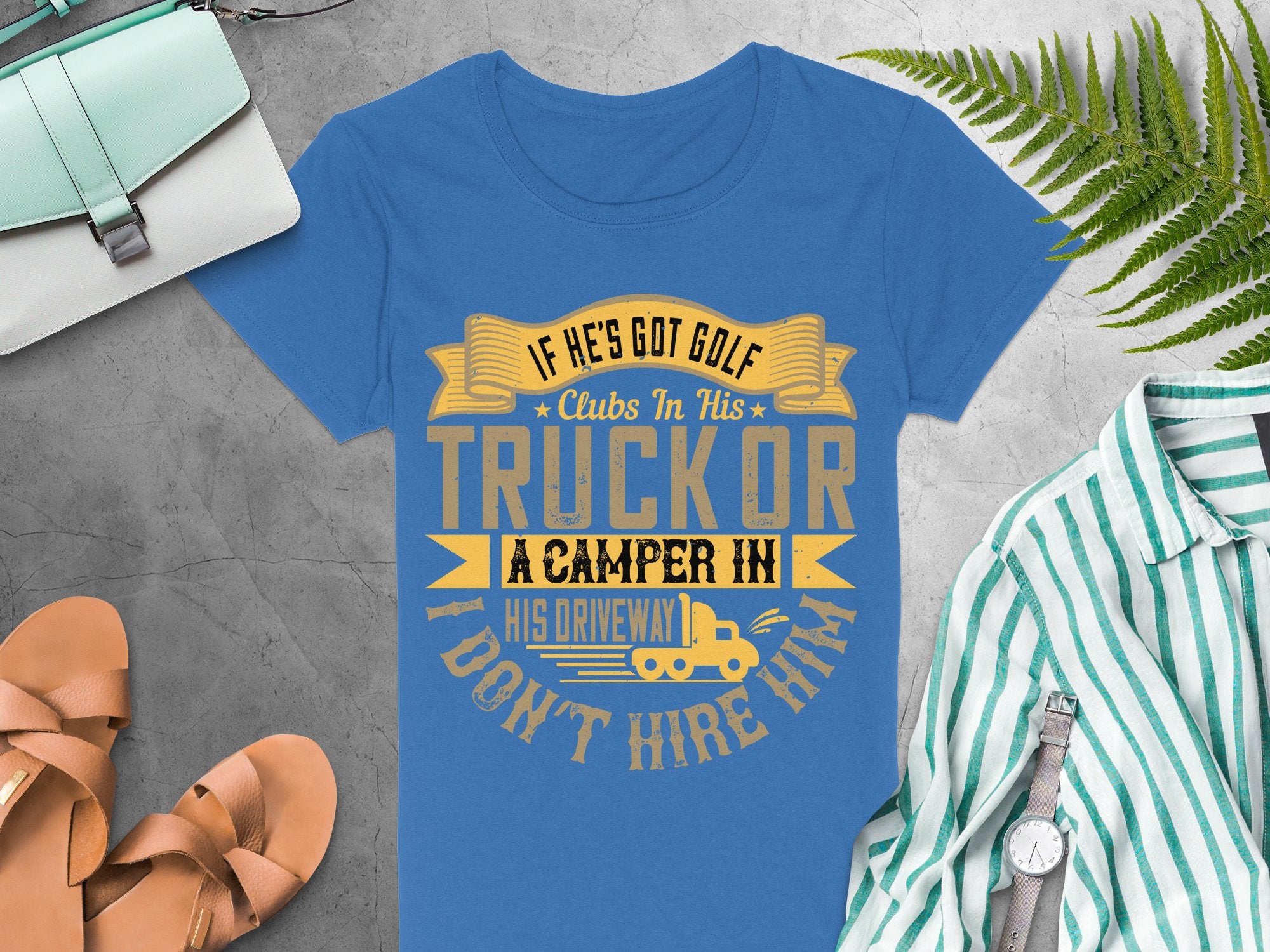 Garment Graphics offers a womens blue T-shirt with a quirky design that reads: If hes got golf clubs in his truck or a camper in his driveway, I dont hire him. Ideal for fans of funny golf graphic tees, its photographed among accessories on a gray surface.