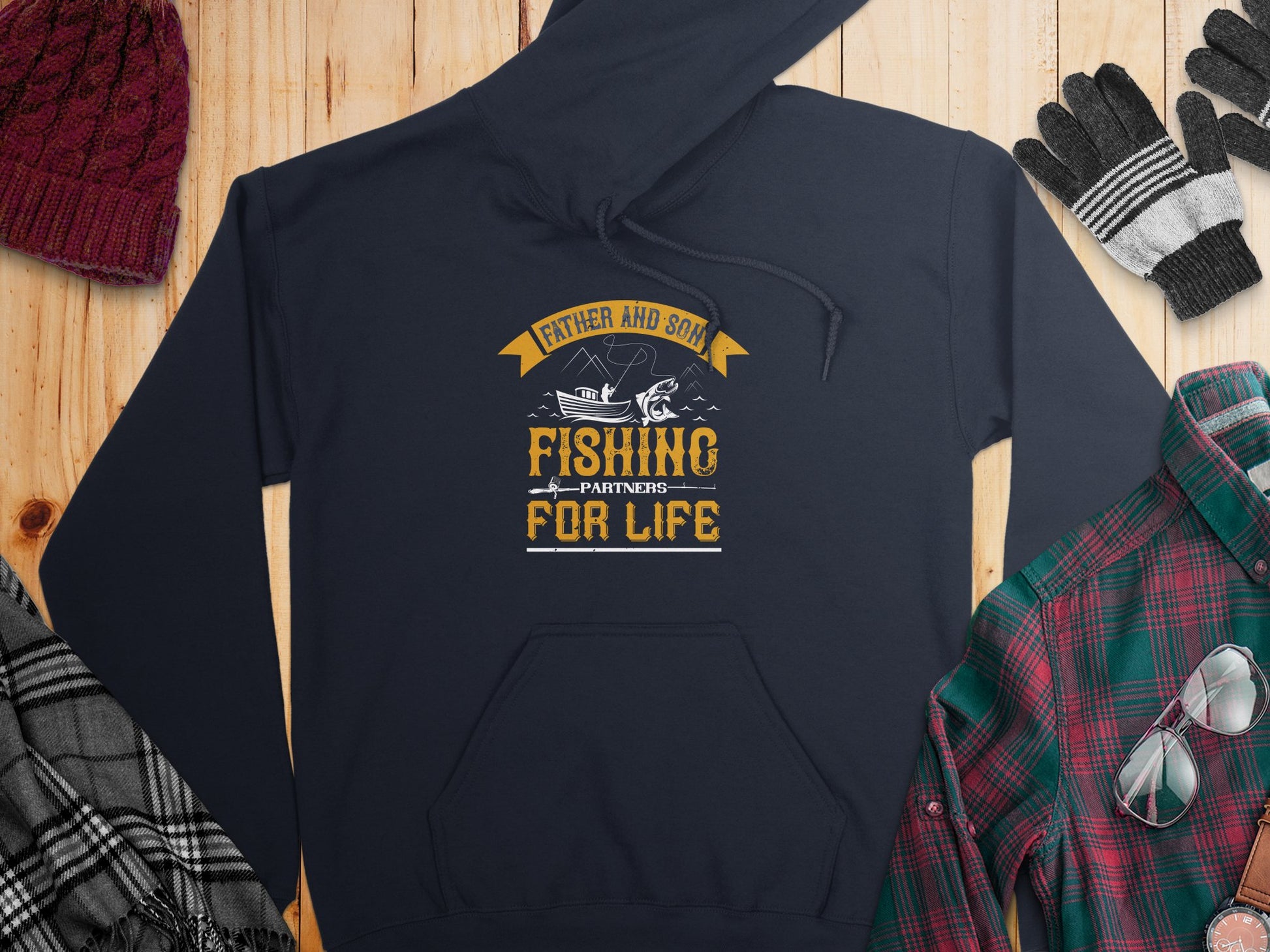 The Garment Graphics navy blue fishing hoodie features Father and Son, Fishing Partners for Life in yellow and white along with a fish and rod design. Ideal for fishing fans, the hoodie is styled on wood with a plaid shirt, gloves, beanie, and scarf.