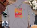 Garment Graphics offers a gray hoodie with Born to be a Musician in orange and red, featuring a musician with guitar and drum set. Ideal for music lovers, it sits on wood surrounded by a camera, binoculars, leather goods, and climbing carabiner.