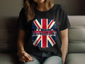 A person relaxes on a sofa in a 100% cotton black t-shirt by Garment Graphics, showcasing a faded Union Jack flag design. The flag boldly states, DONT BLAME ME, I Didnt Vote For Him.