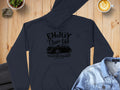 A dark, classic-fit hoodie by Garment Graphics with Enjoy Classic Car and a vintage car design lies on a wooden surface. A cup of latte art adds community spirit, with a stone and folded blue denim shirt nearby.