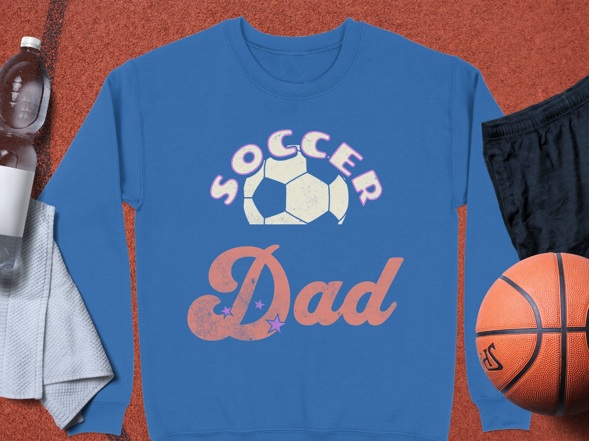The blue Soccer Dad sweatshirt by Garment Graphics showcases a soccer ball design. Its paired with a basketball, black shorts, towel, water bottle, and a glimpse of a red sports court. This stylish unisex sweatshirt is ideal for sports enthusiasts.