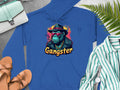 A Garment Graphics blue hoodie features a hipster urban style with an illustrated gangster ape in a hat, sunglasses, and necklace. The word Gangster appears below, surrounded by accessories like a purse, sandals, watch, and striped shirt on a gray background.