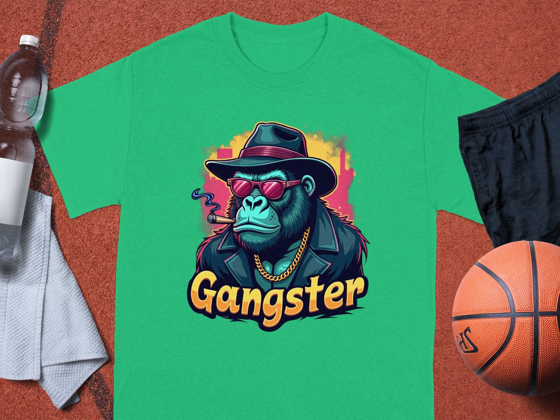 The Garment Graphics Gangster Gorilla T-Shirt features a classic fit with an urban-style gorilla graphic, complete with hat, sunglasses, and gold chain. Gangster is printed below. It is set against an orange backdrop alongside basketball gear and a water bottle.