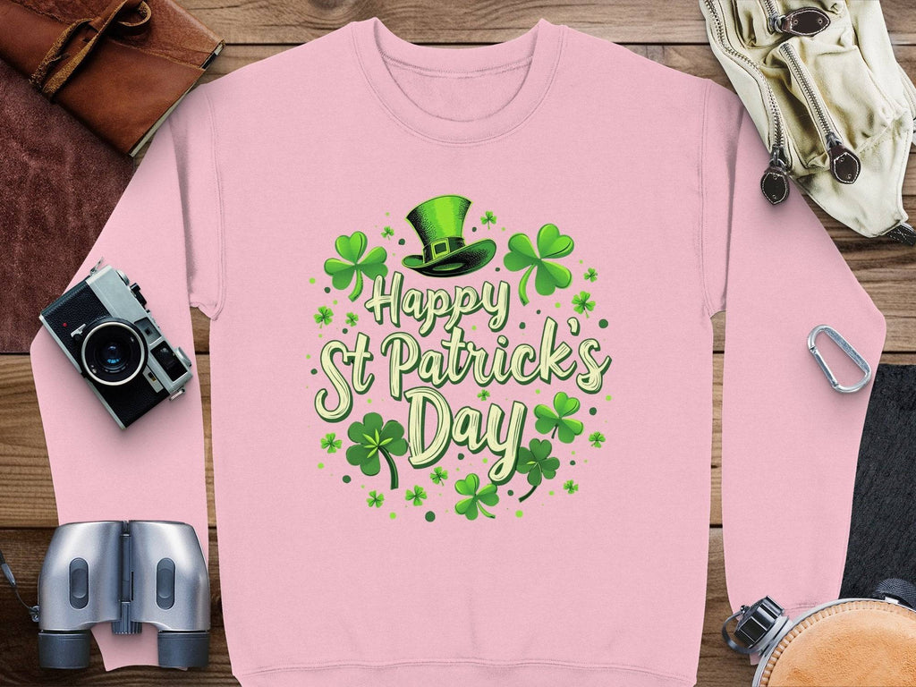 A flat lay showcases a Garment Graphics pink shamrock sweatshirt with Happy St Patricks Day alongside festive accessories: a green leprechaun hat, camera, leather gloves, binoculars, carabiners, and rolled-up map on a wooden surface.