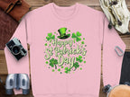 A flat lay showcases a Garment Graphics pink shamrock sweatshirt with Happy St Patricks Day alongside festive accessories: a green leprechaun hat, camera, leather gloves, binoculars, carabiners, and rolled-up map on a wooden surface.