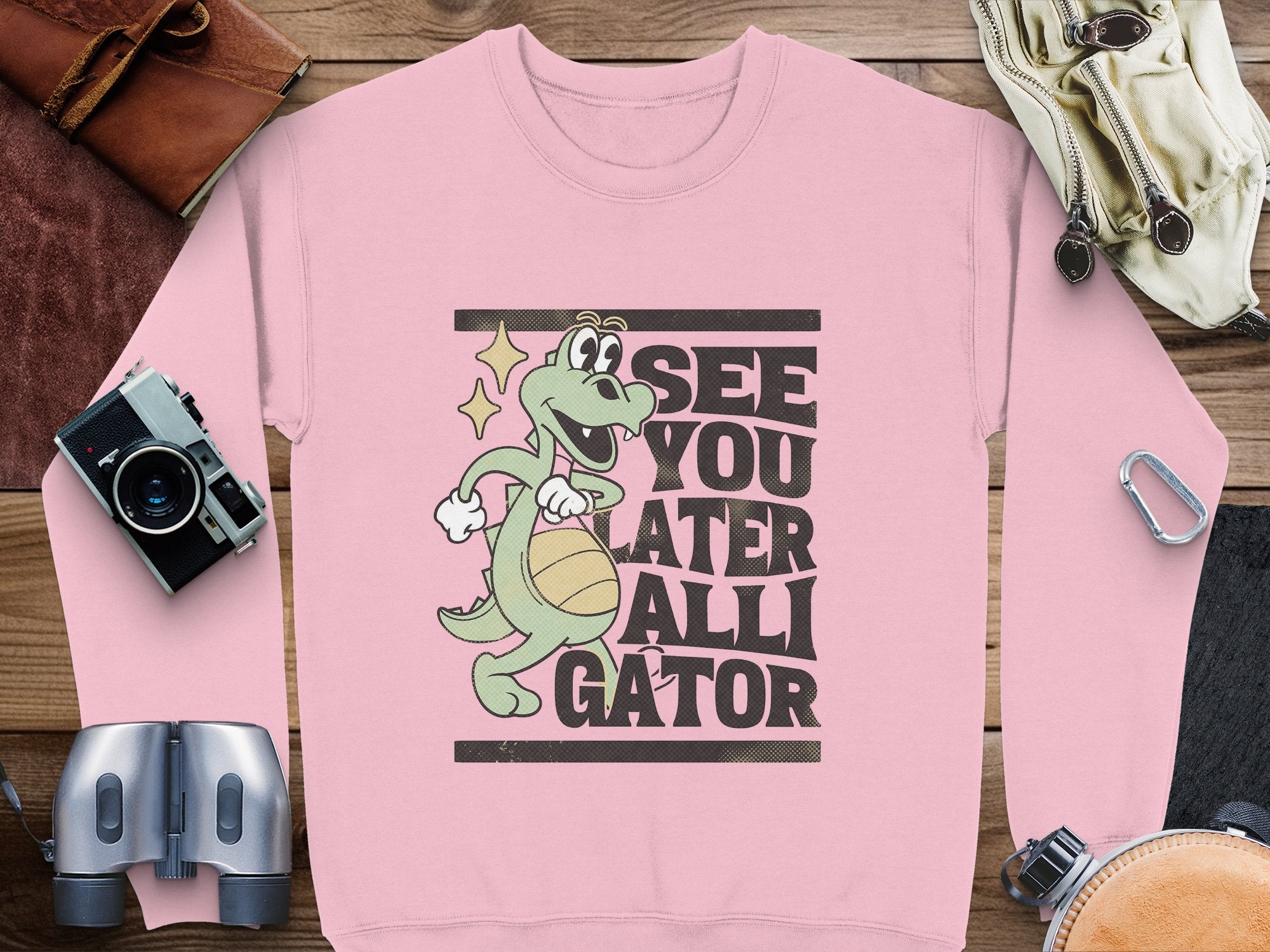 A fun pink sweatshirt from Garment Graphics features a cartoon alligator with a basketball and the phrase See You Later Alligator, creatively arranged on a wooden surface alongside accessories like a camera, binoculars, and leather goods.