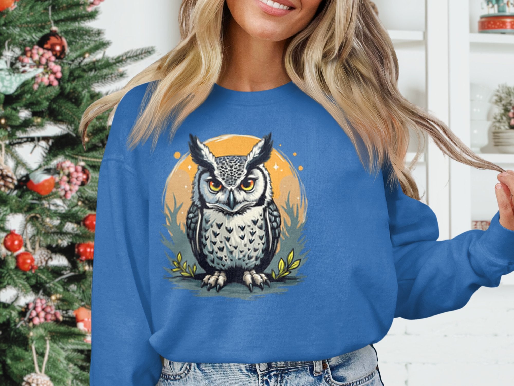 A person stands indoors near a decorated Christmas tree wearing a cozy blue unisex sweatshirt from Garment Graphics. The sweatshirt features an owl with large eyes and folded wings against an orange circular background, complementing their long, wavy hair.