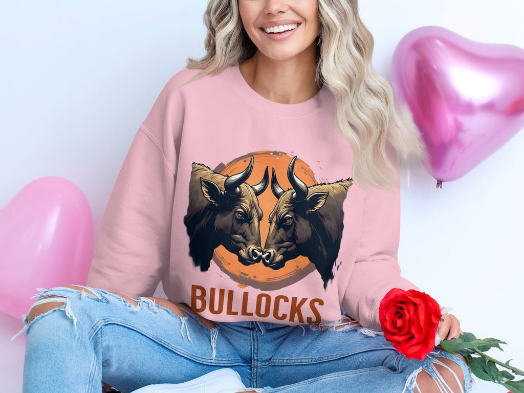 A woman is smiling and holding a red rose while wearing a Garment Graphics loose-fit pink sweatshirt with a bold bullocks graphic featuring two illustrated bulls. She pairs this with ripped jeans and sits near pink heart-shaped balloons.