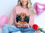 A woman is smiling and holding a red rose while wearing a Garment Graphics loose-fit pink sweatshirt with a bold bullocks graphic featuring two illustrated bulls. She pairs this with ripped jeans and sits near pink heart-shaped balloons.