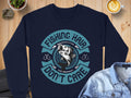 A Garment Graphics navy fishing sweatshirt with Fishing Hair Dont Care and fish graphic sits on a wooden table. Next to it, a denim jacket lies near a potted plant, rocks, and a steaming latte in a white cup—ideal for any fishing enthusiast!.