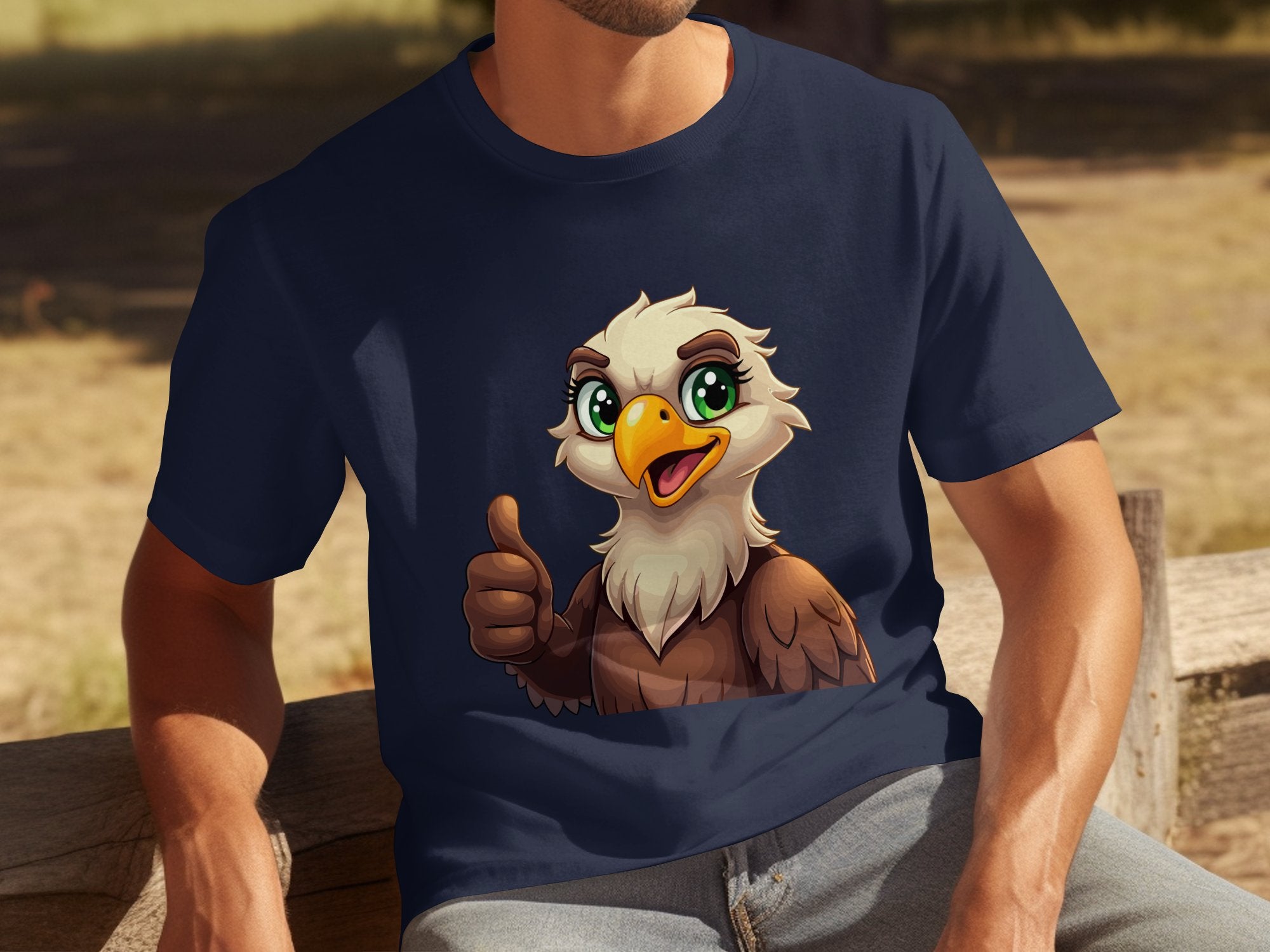 A person sits on a wooden bench, wearing a Garment Graphics navy Eagle T-shirt with a cartoon eagle giving a thumbs-up. This fun casual piece complements the blurred outdoor setting of grass and trees, offering both style and comfort with its classic fit.