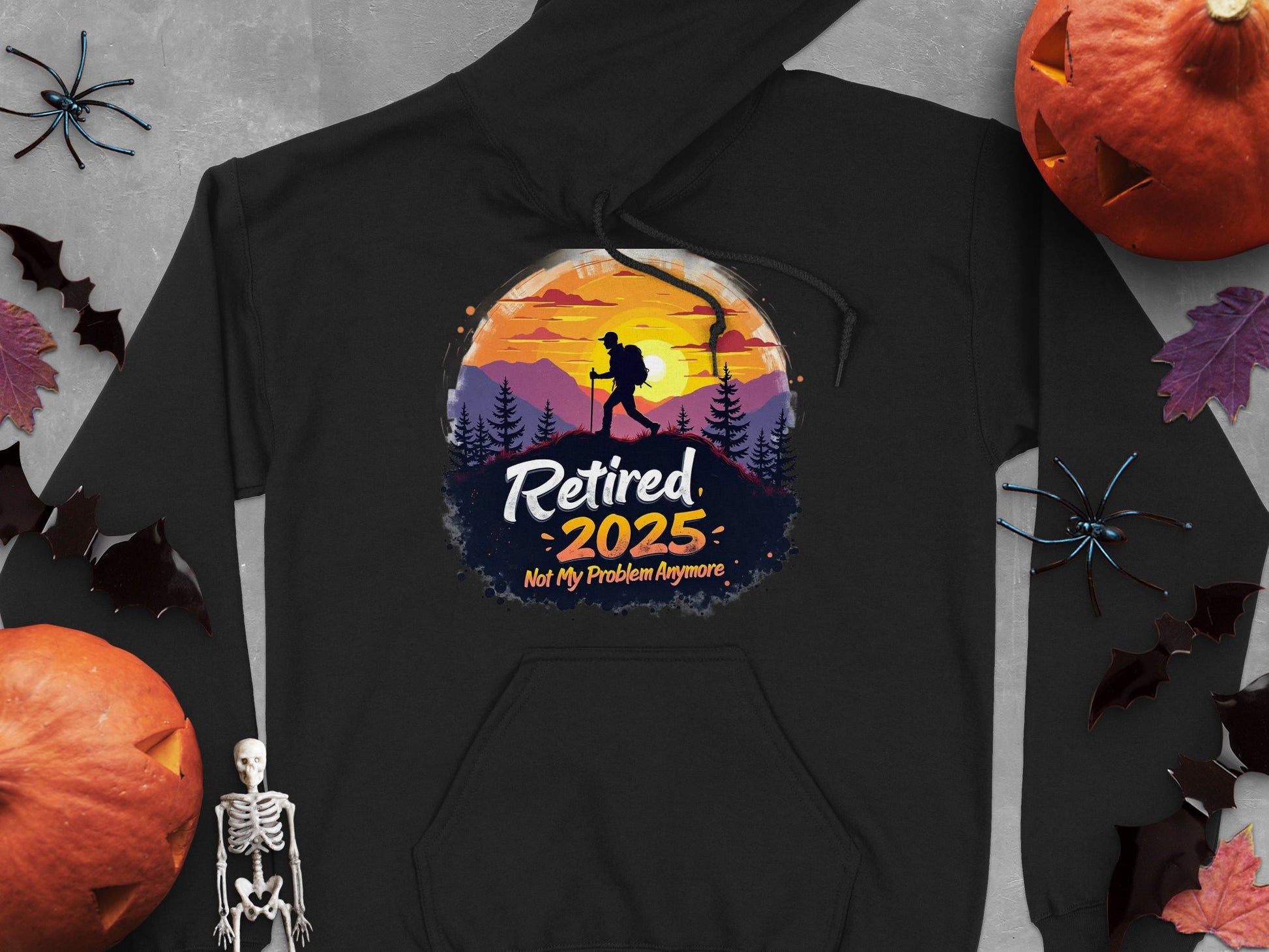 The Garment Graphics black hiking hoodie showcases a sunset mountain design with trees and a hiker, plus the message Retired 2025: Not My Problem Anymore, surrounded by pumpkins, a small skeleton, decorative leaves, and bats.