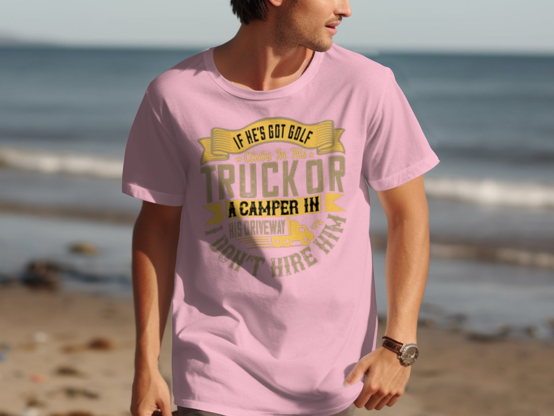 A person in a light pink Garment Graphics t-shirt with golf and camping designs stands on a beach; the serene ocean stretches behind them. The shirt artfully depicts a truck and camper in a driveway while they gaze away from the camera.