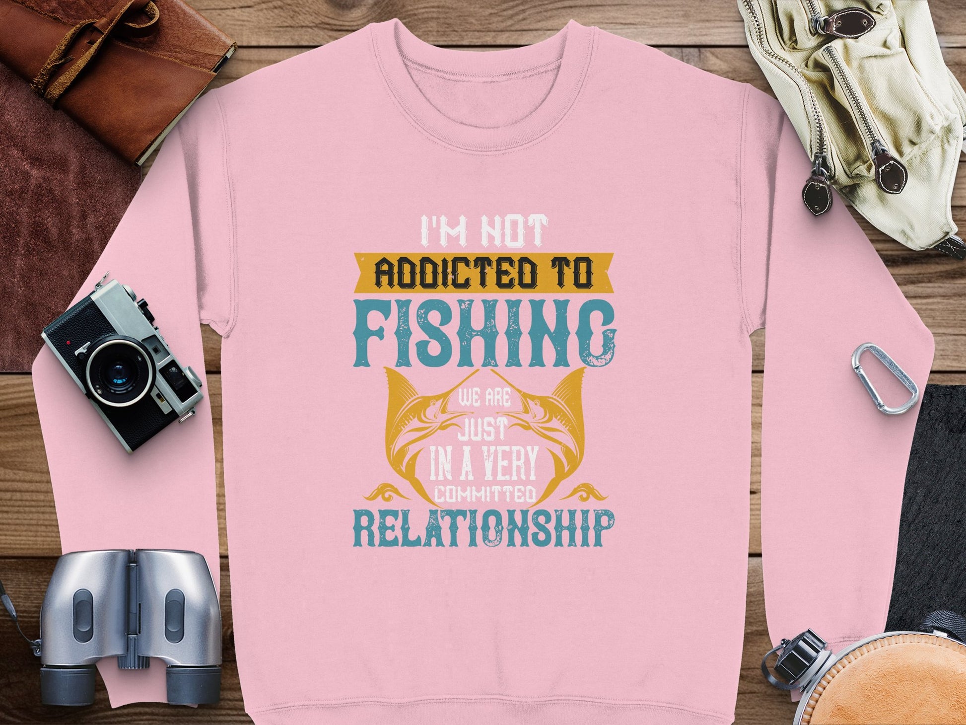 A pink unisex sweatshirt from Garment Graphics, featuring Im not addicted to fishing, we are just in a very committed relationship and fish graphics, lies on a wooden surface. Nearby items include a camera, binoculars, gloves, and a leather pouch.