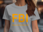 Someone is wearing a Garment Graphics FBI Female Boob Inspector T-shirt, intended as a gag gift. Bold large yellow FBI letters are featured with playful text below. This quirky womens tee makes an ideal gift for the bold-hearted, with the background casually blurred.