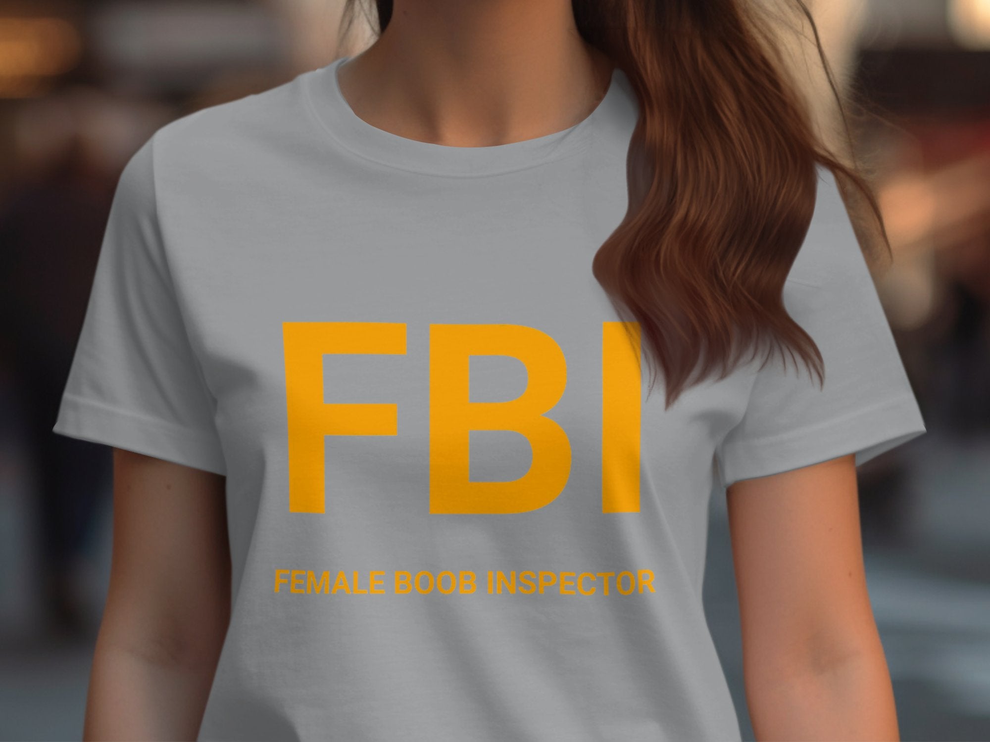Someone is wearing a Garment Graphics FBI Female Boob Inspector T-shirt, intended as a gag gift. Bold large yellow FBI letters are featured with playful text below. This quirky womens tee makes an ideal gift for the bold-hearted, with the background casually blurred.