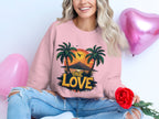 A person with wavy hair wears a Garment Graphics pink sweatshirt featuring palm trees and One Love Sunset design, holding a red rose while sitting beside heart-shaped balloons in pink and purple, perfectly complementing their ripped jeans.